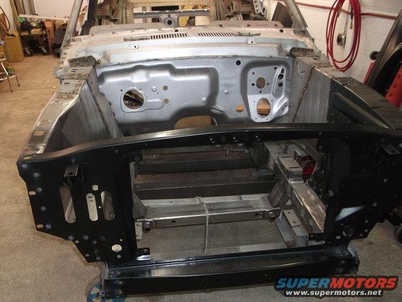 Car chassis on a dolly, showing the engine bay and front frame. The body is partially stripped