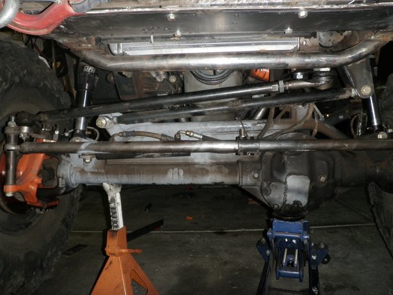 Underside view of a vehicle's steering linkage, tie rods, and front axle assembly, supported by jack stands