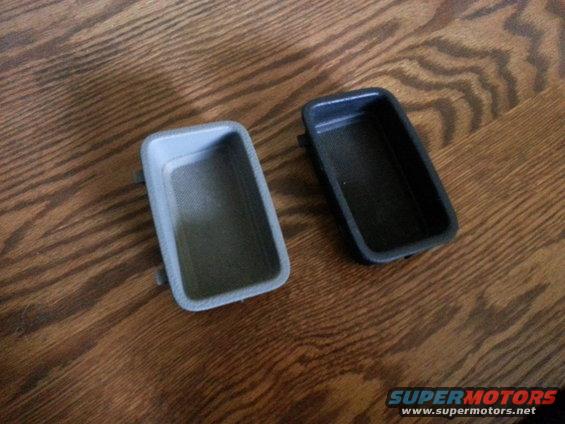 Two rectangular plastic car interior trim pieces, one gray and one black, on a wooden surface