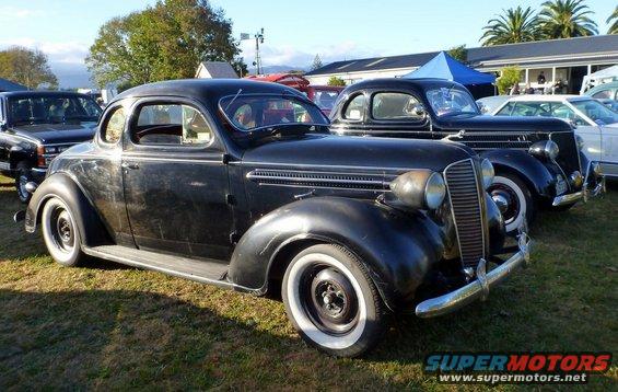 Black vintage coupe with whitewall tires parked on grass, another similar car behind it
