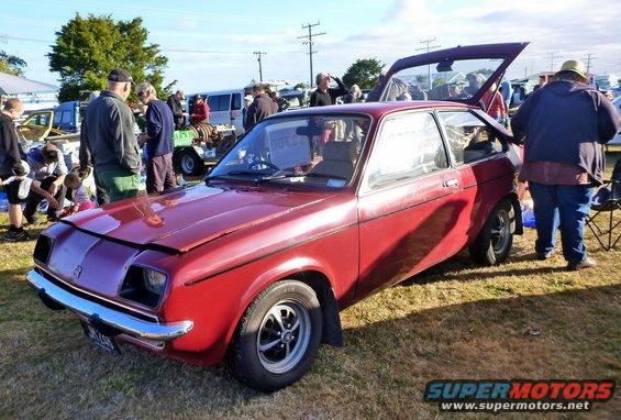 Red hatchback with hood open and trunk open, parked on grass at an outdoor event with people around