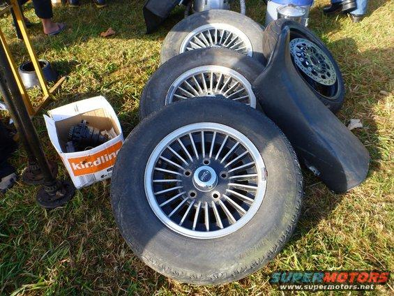 Several car wheels and a black fender are on the grass next to a box of parts