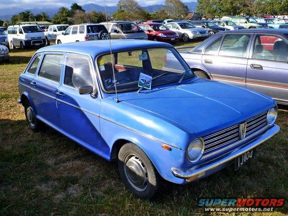 Blue hatchback with chrome grille and license plate IJ804 parked on grass, other cars in background