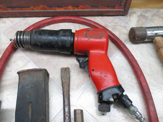 Red air hammer with black barrel and spring, next to chisels and a hammer