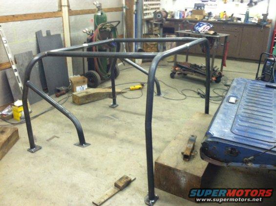 Custom roll cage fabricated from steel tubing, set up in a workshop with tools and a blue vehicle tailgate visible