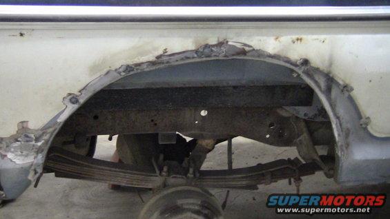 White vehicle's rusted rear wheel well and leaf spring suspension, showing significant body damage