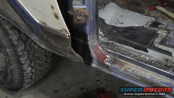 Vehicle door frame with rust and repair work, showing a tire and part of the body. The metal is exposed and welded