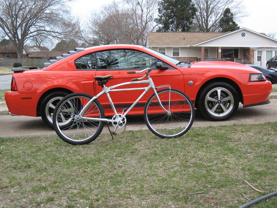 Red Ford Mustang coupe with louvers on rear window, parked next to a silver bicycle