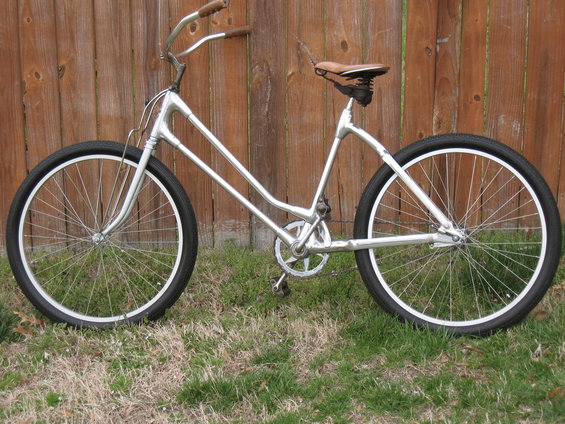 Silver cruiser bicycle with brown handlebars and seat, parked on grass in front of a wooden fence