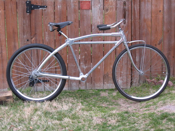 Custom silver cruiser bicycle with wide tires and a spring seat, parked in front of a wooden fence