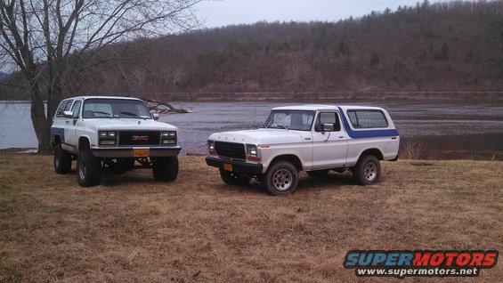 White Ford Bronco and white GMC SUV parked on grassy shore near water