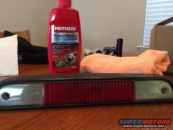 Car tail light assembly with red and clear sections, Mothers PowerPlastic 4 Lights bottle, orange microfiber cloth, and paper towels on a wooden table