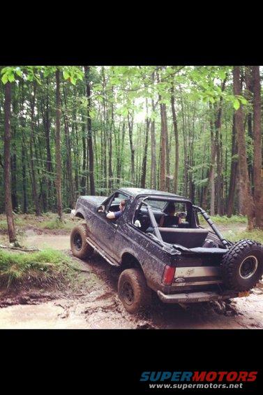 Ford Bronco driving through a muddy trail in a forest. It has a spare tire mounted on the back