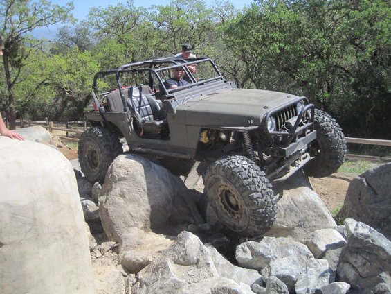 Modified Jeep Wrangler navigating large rocks on a trail. The vehicle has a roll cage and large off-road tires