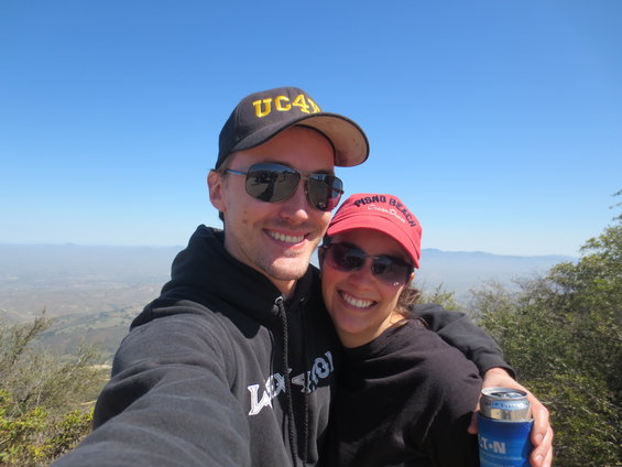 Two smiling people in hats and sunglasses take a selfie with a hazy landscape behind them