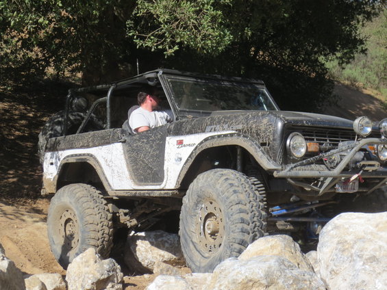 Mud-splattered first-generation Ford Bronco with off-road modifications navigating rocks