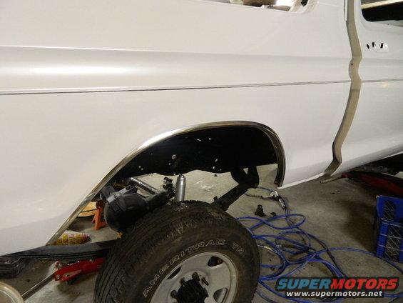 White pickup truck's rear wheel well area, showing suspension and tire. Some blue cords are on the floor