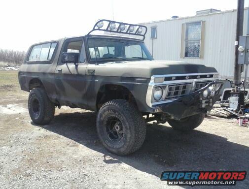 Ford Bronco with camo paint, roof rack, and aftermarket bumper parked outside a trailer