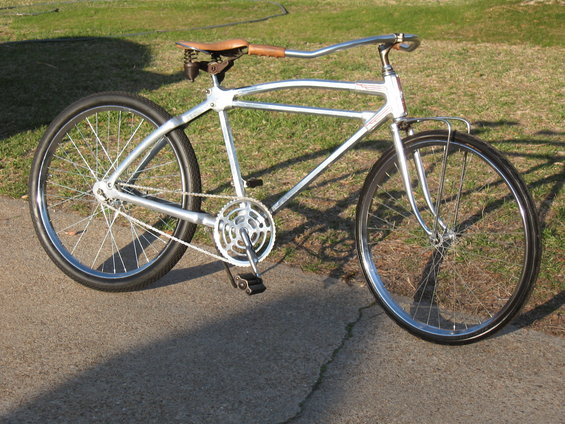 Silver custom bicycle with leather seat and chrome handlebars, parked on pavement with grass in background