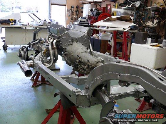 Car frame on jack stands with engine and transmission installed. Exhaust pipes visible