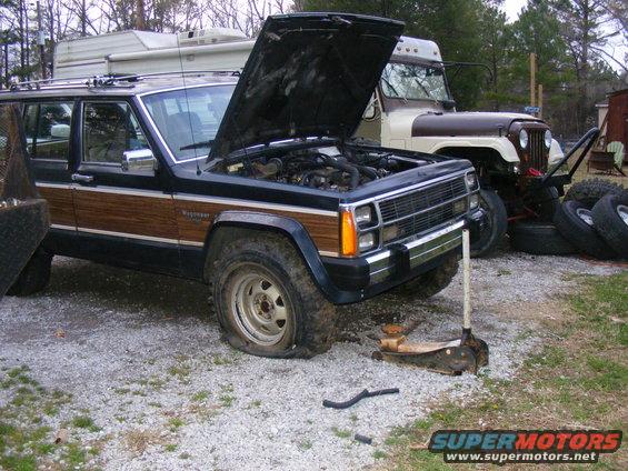 Black Jeep Grand Wagoneer with wood paneling, hood open, and flat front tire