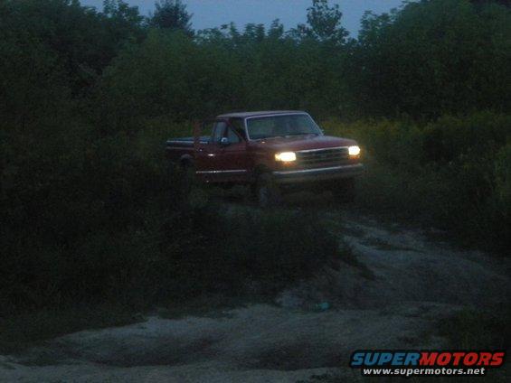 Red pickup truck driving on a dirt path at dusk, headlights on