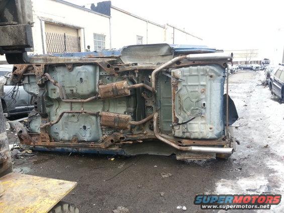 Blue car chassis upside down, showing rusted exhaust pipes and undercarriage. A forklift arm is visible on the left