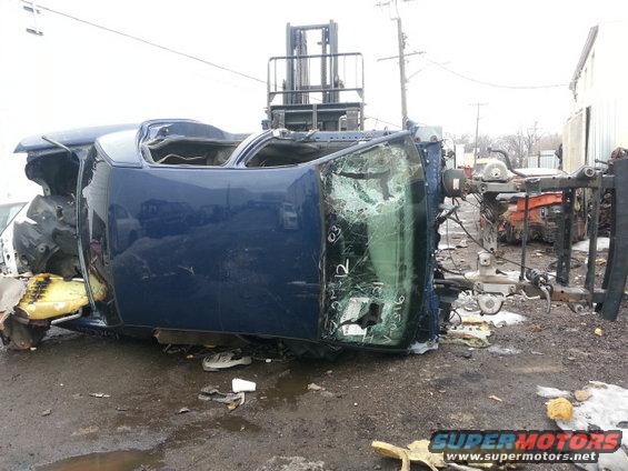 Wrecked blue car body, possibly a sedan, overturned and being moved by a forklift. Windshield is broken