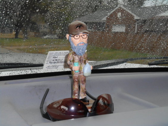 Bobblehead figure of a man with glasses and beard holding a blue bottle and a white bag, resting on a dashboard with sunglasses