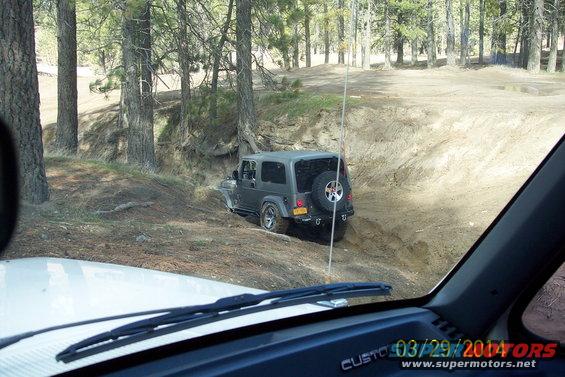 Dark gray Jeep Wrangler driving down a dirt path in a wooded area, seen from inside another vehicle