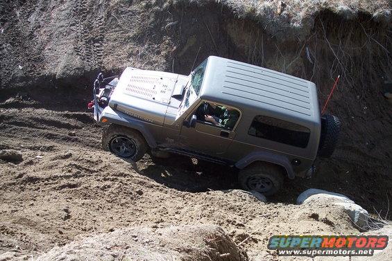 Gray Jeep Wrangler Rubicon off-roading on a dirt hill, front-left tire stuck in mud