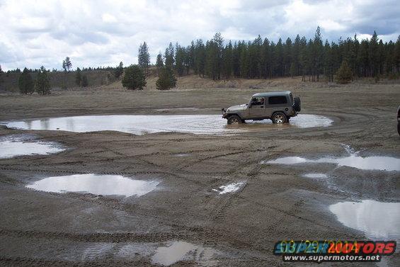 Gray Jeep Wrangler driving through a mud puddle in a field with trees in the background. Another vehicle is partially visible on the right