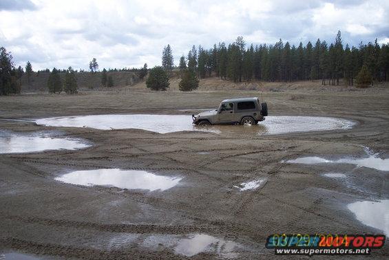 Gray Jeep Wrangler stuck in a muddy puddle with tire tracks in the foreground