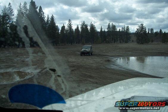 Jeep driving on a muddy track near a body of water, seen from inside another vehicle