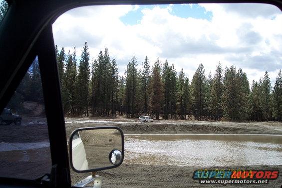 White SUV on dirt road, trees and water in background. Another vehicle visible to the left