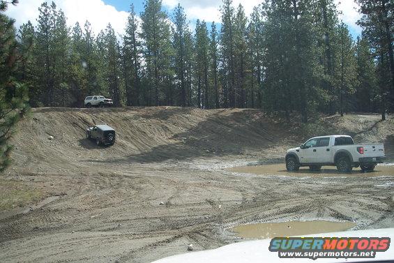 White pickup truck and two SUVs on a muddy off-road course with pine trees in the background