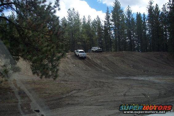 Two trucks driving up a dirt hill in a wooded area. One is white, the other is white