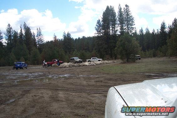 Several off-road vehicles, including a blue SUV and red truck, are in a muddy area with trees in the background