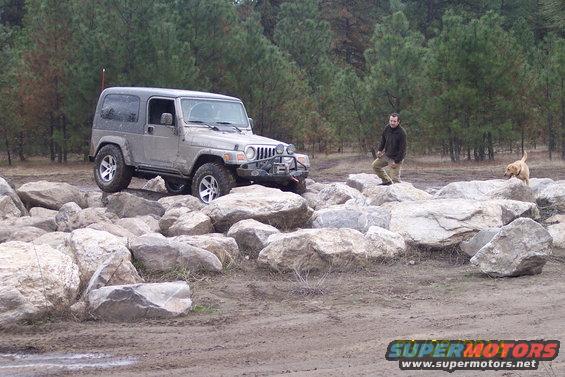 Silver Jeep Wrangler on rocks, front-left view, with a man and dog in background