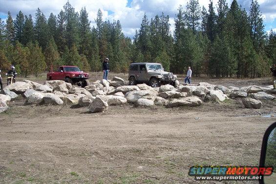 Red pickup truck and silver Jeep on rocky terrain with trees in background