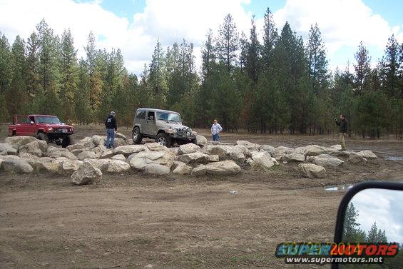 Red pickup truck and silver Jeep Wrangler on a rocky trail with people standing nearby. Trees and sky in background