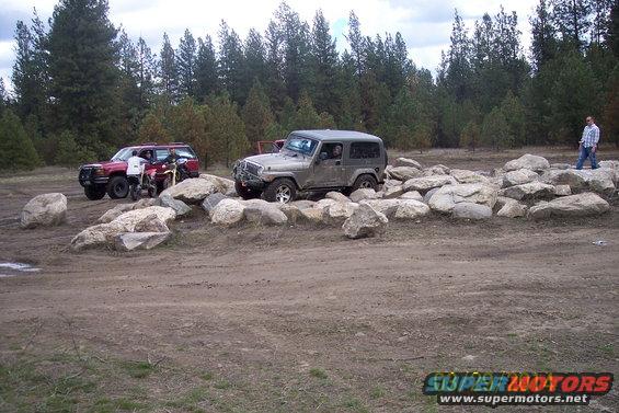 Gray Jeep Wrangler on rocks, red SUV and dirt bike nearby