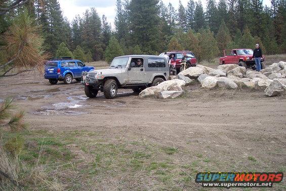 Several off-road vehicles, including a silver Jeep, a blue SUV, and a red pickup truck, navigate a rocky, muddy track