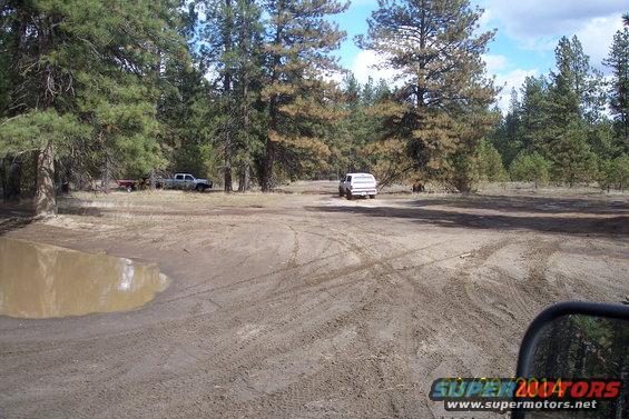 Off-road scene with a white van and a pickup truck parked near trees, with a muddy puddle in the foreground