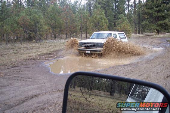 White SUV splashing through a mud puddle on a dirt road, seen from a side mirror