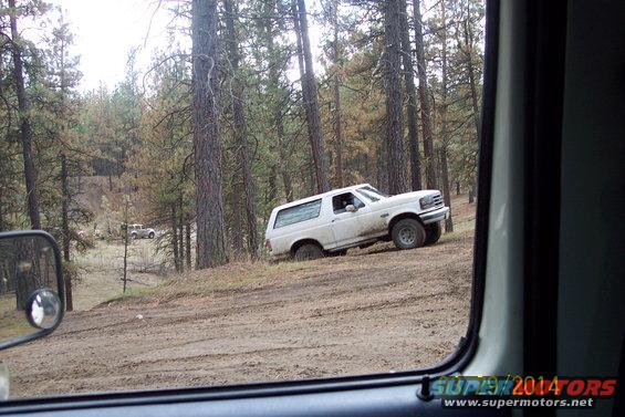 White Ford Bronco stuck in mud on a dirt road, viewed from inside another vehicle