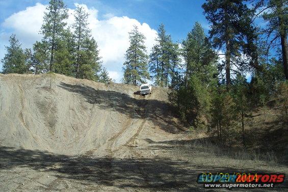 White SUV on a steep dirt hill with tire tracks, pine trees and blue sky