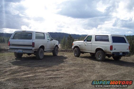 Two white Ford Broncos parked on a dirt road, one facing rearward, the other 3/4 front. The Bronco on the right appears to be a 1992 model