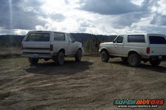 Two white 1990 Ford Broncos parked on a dirt road with trees and cloudy sky in background