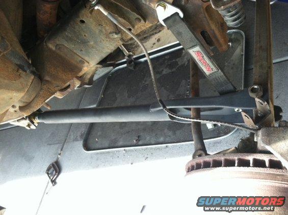 Close-up of a vehicle's suspension with a black strap and metal components visible. Supermotors.net logo at bottom right